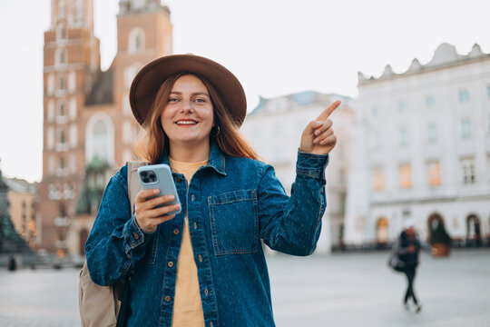 Attractive young female tourist is exploring new city. Redhead girl pointing finger and holding a smart phone on Market Square in Krakow. Traveling in summer. St. Marys Basilica. Vacation concept
