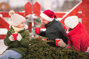 Family drinking tea to keep warm on a sleigh with a Christmas tree in a snowy winter outside.