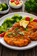 Crispy breaded fried chicken cutlet with boiled broccoli and fresh vegetables on wooden table
