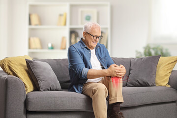 Mature man in pain sitting on a sofa at home and holding his inflamed knee