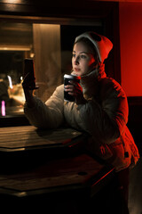 Model blonde with tails with cup of coffee near coffee shop with red and yellow light looking in smartphone. Girl weared white dress, jacket and hat posing near cafe in winter night