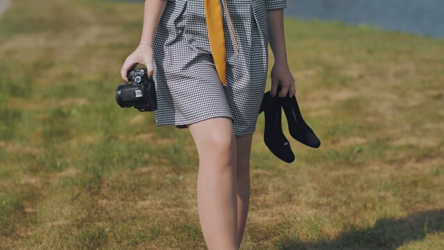 A barefoot girl photographer walks through a meadow with a camera and shoes.