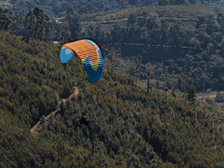 paraglider with the colorful sail, skirting a tree-covered hill while heading towards the valley.