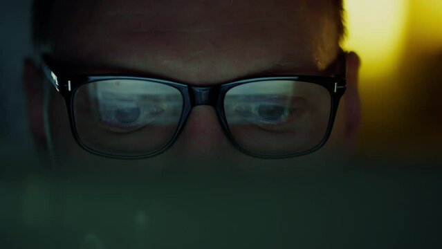 Close-up of the eyes of a businessman working at night studying information analyzing data sitting at a portable laptop. Information flickers in the reflections of the glasses of his eyes