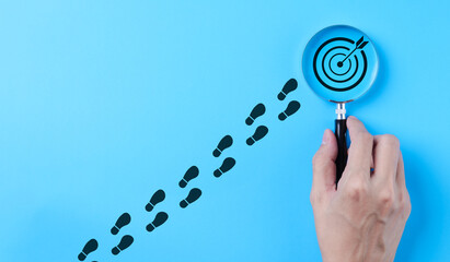 Hand holding magnifying glass focusing on target goal icon on blue background. Business achievement...