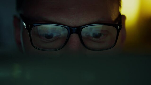 In a close-up, the eyes of a businessman working hard at night closely study information and analyze data from a portable laptop. Data reflections flicker in his glasses