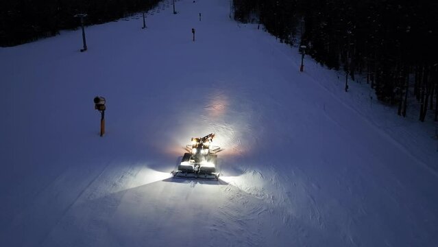 Aerial view of working snowcat on the snowy alpine ski slope at winter night in Kranjska Gora, Slovenia. Top drone view of snowcat grooming snow, ski resort, lights at dusk. Snowplough machine
