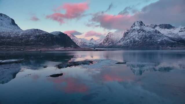 Aerial view of sea, snowy mountains, rorbuer in winter at sunset. Lofoten islands, Norway. Top drone view of fjord, house, reflection in water, stones, rocks, blue sky with pink clouds at dusk. Lake