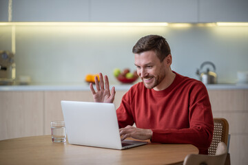 Cheerful man has online business meeting video call sitting on kitchen at home waving hand greeting...