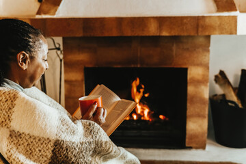 Senior African woman reading a book while warming by cozy fireplace in winter day. Relax and...