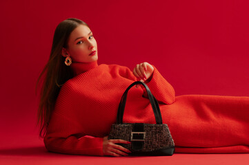 Fashionable confident woman wearing trendy red knitted turtleneck dress, golden earrings, holding baguette bag, posing, laying on red background. Studio fashion portrait. Copy, empty space for text - obrazy, fototapety, plakaty