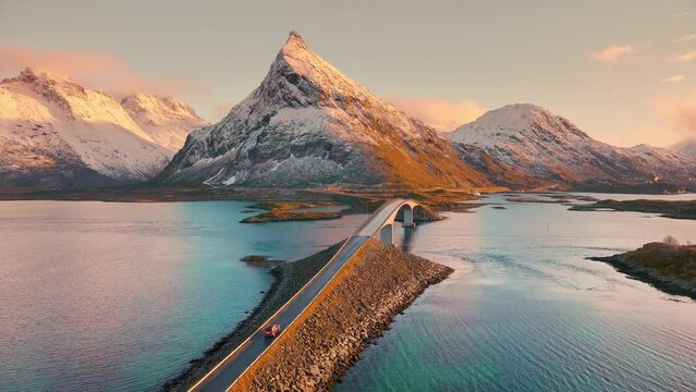 Aerial view of Fredvang bridge, car, sea and snowy mountains at sunset in Lofoten Islands, Norway. Landscape with beautiful road, water, rocks in snow, sky with pink clouds. Top view from drone