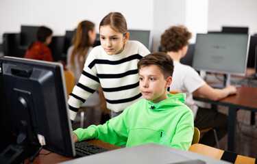 Teenager boy using computer in classroom to help his classmate, young girl, to solve problem.