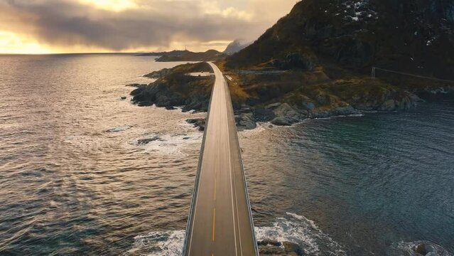 Aerial view of bridge, sea with waves and snowy mountains at sunset in winter. Lofoten Islands, Norway. Top drone view of beautiful road, water, rocks, sky with clouds and golden sunlight. Transport