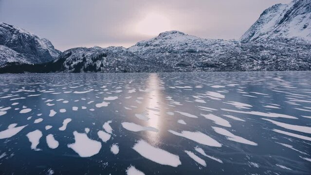 Aerial view from low flying drone of frozen lake in snowy mountains in winter at sunset. Lofoten islands, Norway. Top view of frosted river, reflection on ice, snowy rocks in fog, sky. Cloudy weather