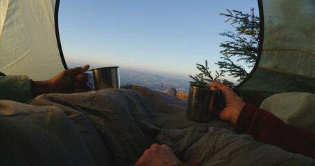 Multiethnic tourist couple sit in tent on mountain hill: They talk, clink cups and drink tea. Two travelers rest during adventure vacation. Romantic backpacker family warm up and admire the scenery.