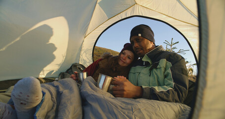Multiethnic backpacker couple sit in tent on mountain hill: They hug and drink tea. Two travelers stopped to rest during adventure vacation. Romantic hiker family admire the nature. Windy weather.