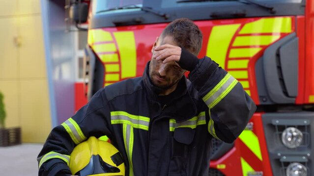 Tired fireman in protective uniform looking at camera while standing near fire engine on station