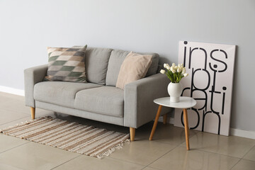 Cozy sofa with cushions and tulip flowers in vase on table near grey wall