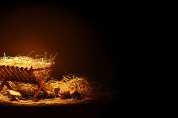 Manger with hay and stones on wooden table against dark background - obrazy, fototapety, plakaty