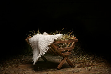 Manger with cloth and hay on wooden table against dark background - obrazy, fototapety, plakaty