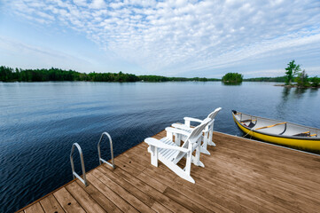 Two white Adirondack chairs on a wooden dock face a calm summer lake. A tied yellow canoe adds to...