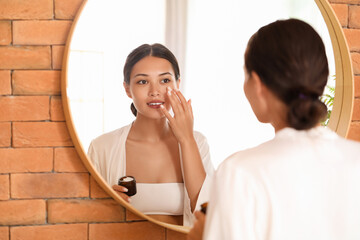 Beautiful Asian woman applying facial cream near mirror in bathroom