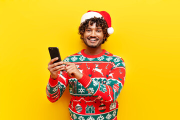 young curly hindu man in new year clothes using smartphone on yellow isolated background