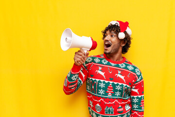 young indian man in new year clothes announces news into megaphone on yellow isolated background