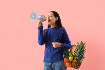 Young woman with shopping basket using megaphone on pink background