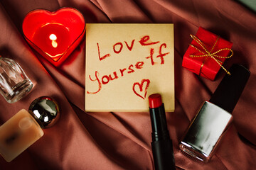 A note with a call to LOVE YOURSELF first drawing with red lipstick on paper for notes on silky pink cloth. A burning heart-shaped red candle, gift box, cosmetic bottles. Happy Valentines Day 2024. - obrazy, fototapety, plakaty