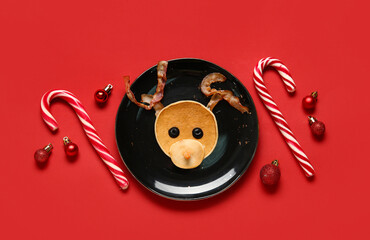 Plate with pancake in shape of reindeer on red background. Christmas celebration - obrazy, fototapety, plakaty