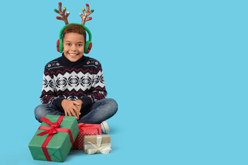 Little African-American boy in winter clothes with deer horns and gifts on blue background