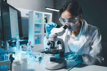 Young scientists in sterile clothing and safety goggles sitting at table conducting research investigations in a medical laboratory using a microscope, Serious concentrated female microbiologist. - obrazy, fototapety, plakaty