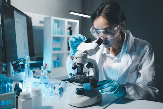 Young scientists in sterile clothing and safety goggles sitting at table conducting research investigations in a medical laboratory using a microscope, Serious concentrated female microbiologist.