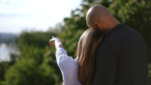 Rear view of a happy couple standing in a spring park. The woman points her hand to the city spread across the river