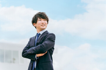 青空とオフィスの前に立つ若いアジア人ビジネスマン（腕組み・見上げる）
 - obrazy, fototapety, plakaty
