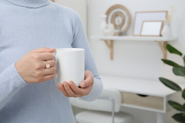 Woman holding white mug at home, closeup and space for text. Mockup for design