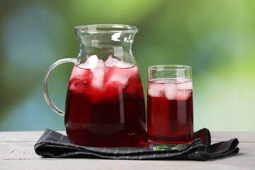 Refreshing hibiscus tea with ice cubes on white wooden table against blurred green background