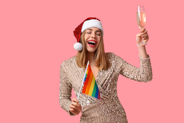 Fototapeta na wymiar Young woman in Santa hat with LGBT flag and champagne on pink background