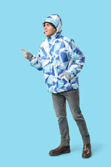 Young man in stylish puffer jacket and gloves pointing at something on blue background