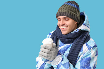 Young man in stylish puffer jacket and scarf with paper cup of coffee on blue background