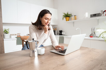 Pretty young woman with geyser coffee maker and cup of espresso using laptop in modern kitchen