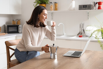 Pretty young woman with geyser coffee maker, cup of espresso and laptop in modern kitchen