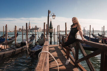 A woman in a black dress poses on a pier in Venice, Italy, with gondolas in the background. The...