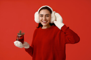 Happy young woman in warm mittens with glass of hot mulled wine on red background