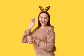 Happy young woman in reindeer horns with cup of hot mulled wine on yellow background