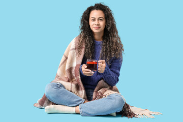 Beautiful young African-American woman with glass cup of hot mulled wine and plaid on blue background