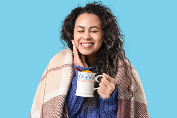 Beautiful young African-American woman with cup of hot mulled wine and plaid on blue background