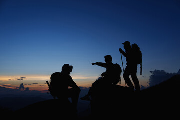 Fototapeta na wymiar Silhouette of Asian teamwork standing raised hands with trekking poles and Backpack and other camping gear on cliff edge on top of rock mountain with at sunset rays over the clouds background, 